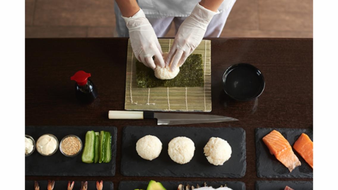 Przygotowanie sushi: ręce w rękawiczkach formują ryż na nori, obok składniki jak łosoś, ogórek i sos sojowy na ciemnym blacie.