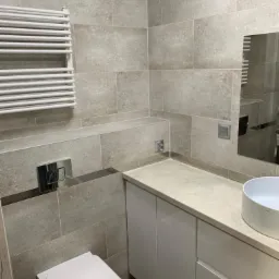 Wykończona łazienka z szarymi płytkami, białymi meblami łazienkowymi, grzejnikiem drabinkowym i wiszącą miską WC.