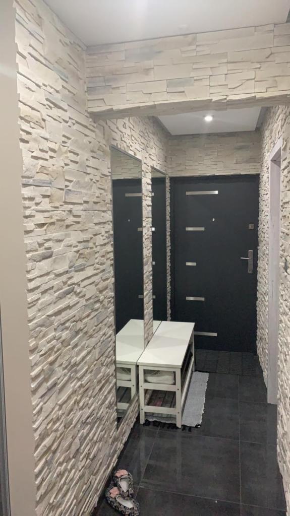 Wąski korytarz z dekoracyjną ścianą z jasnego kamienia, czarnymi drzwiami wejściowymi z poziomymi paskami i lustrem odbijającym białą szafkę na buty. Na podłodze leżą kapcie.
