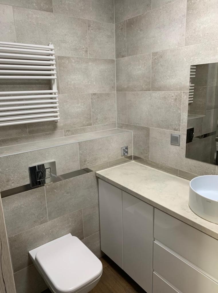 Wykończona łazienka z szarymi płytkami, białymi meblami łazienkowymi, grzejnikiem drabinkowym i wiszącą miską WC.