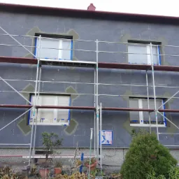 Dom w trakcie ocieplania elewacji, widoczne rusztowania, zabezpieczone okna folią i obróbka szpalet wokół okien.