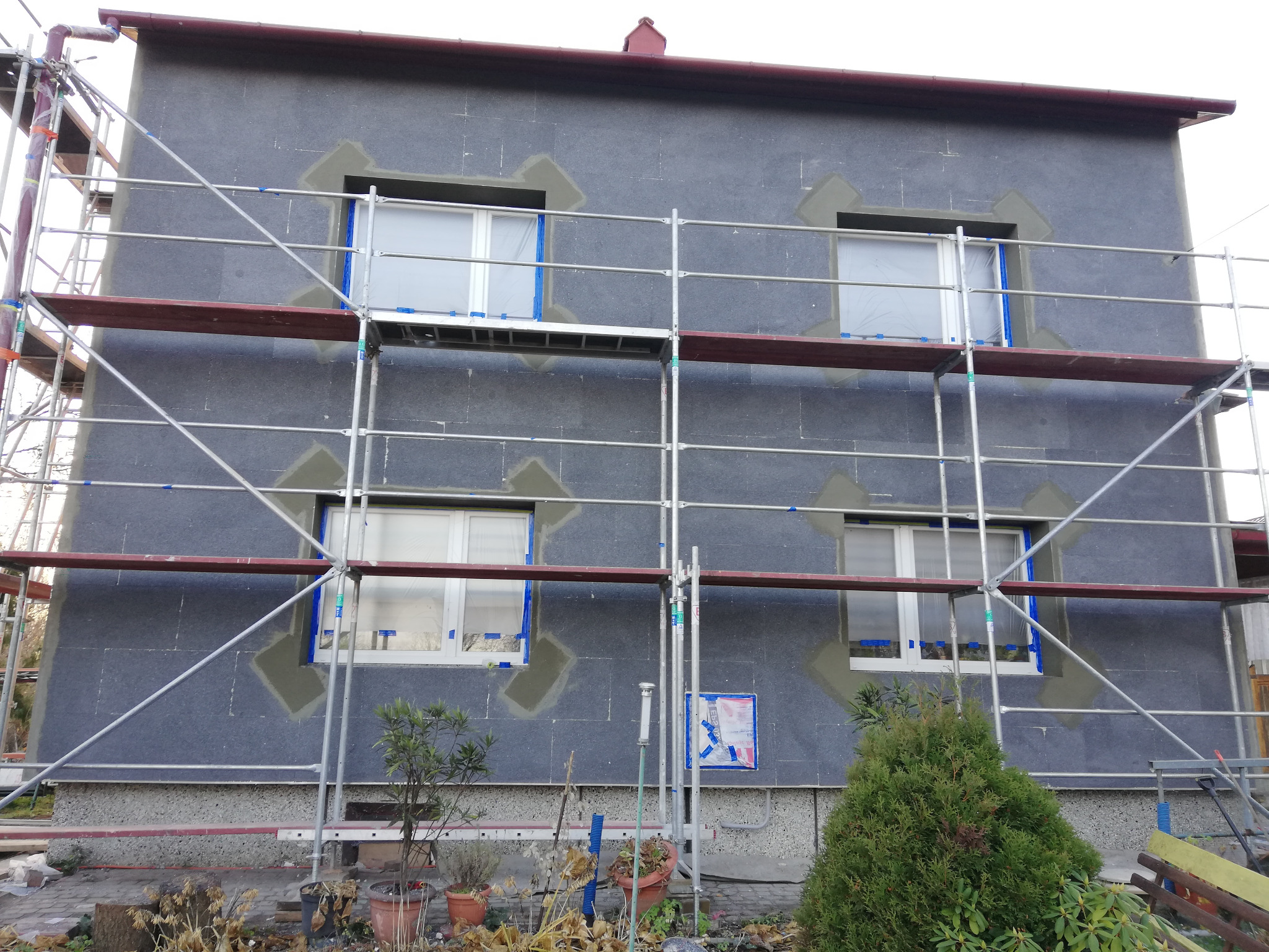 Dom w trakcie ocieplania elewacji, widoczne rusztowania, zabezpieczone okna folią i obróbka szpalet wokół okien.