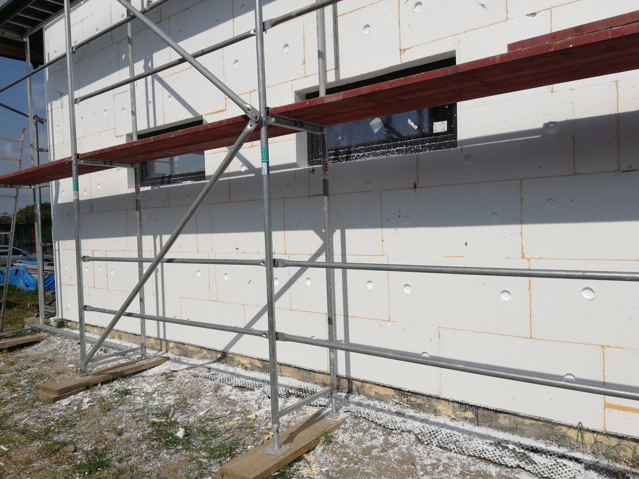 Elewacja budynku w trakcie prac ociepleniowych, widoczne rusztowanie, białe płyty styropianowe z widocznymi otworami, fragment okna z folią ochronną, oraz resztki materiałów na ziemi.