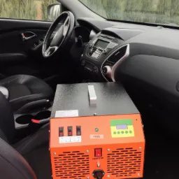 Pomarańczowe urządzenie z napisem PRACA, włącznikami i wyświetlaczem ustawione na skórzanym fotelu samochodowym, wnętrze pojazdu na tle drzew.