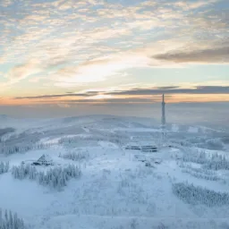 Szeroki kadr na zimowy krajobraz górski z wieżą telekomunikacyjną, budynkami i lasem pokrytym śniegiem, sfotografowany o wschodzie słońca z widocznymi chmurami na niebie.