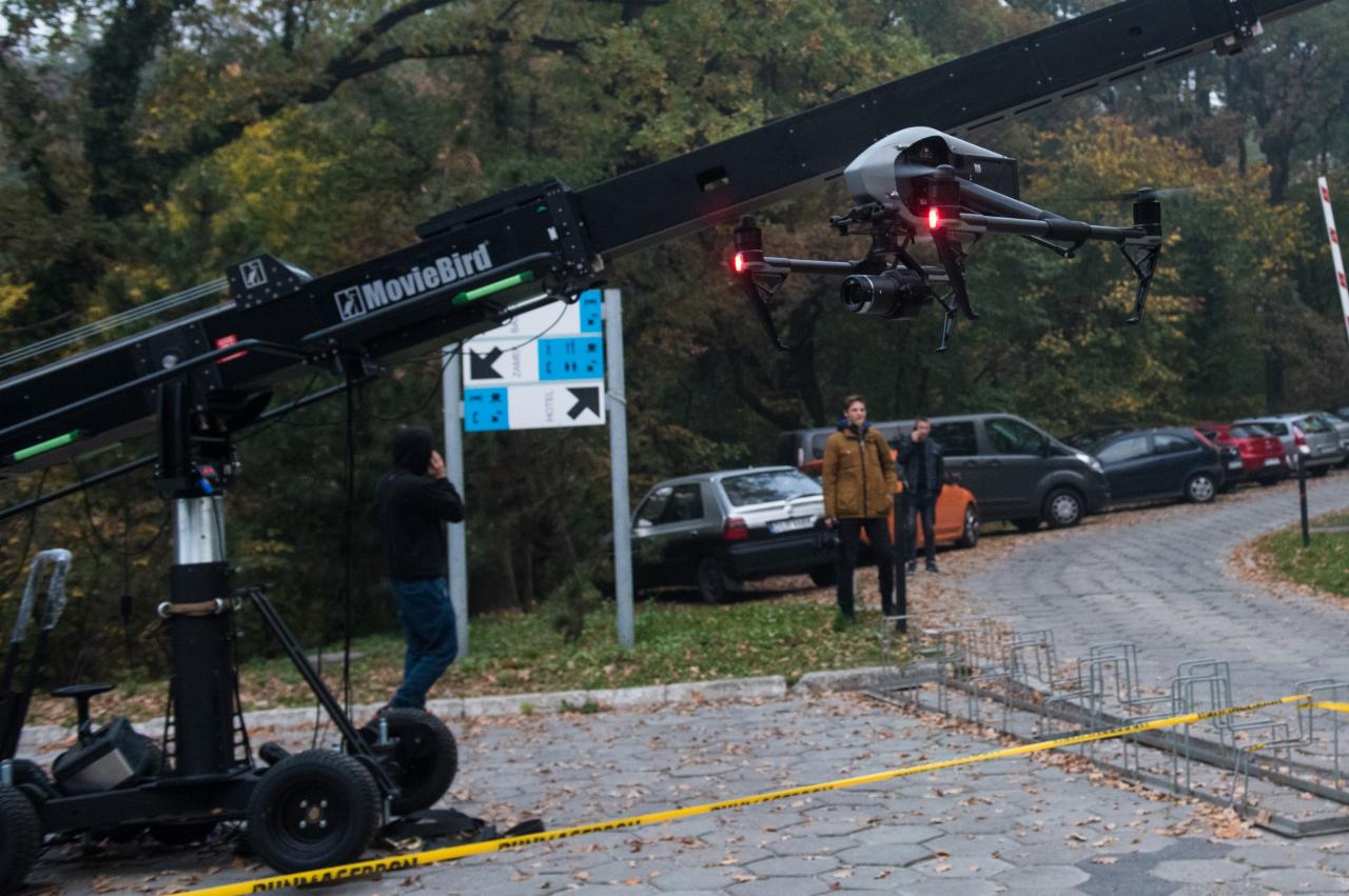 Kamera drona zamontowana na wysięgniku filmowym MovieBird podczas kręcenia ujęć na zewnątrz, w tle zaparkowane samochody i osoby obserwujące.