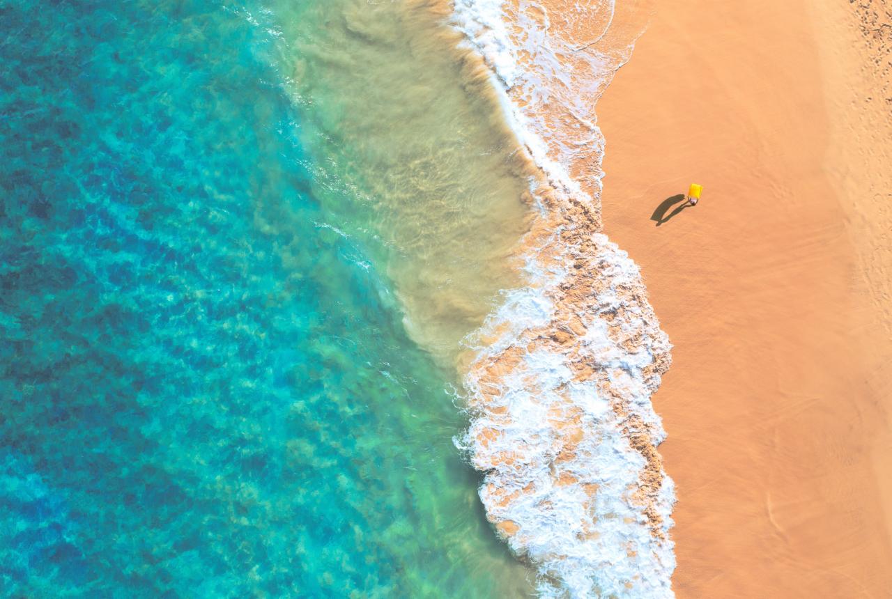 Widok z lotu ptaka na turkusowy ocean i piaszczystą plażę z pojedynczą osobą w żółtym stroju, spacerującą wzdłuż brzegu.