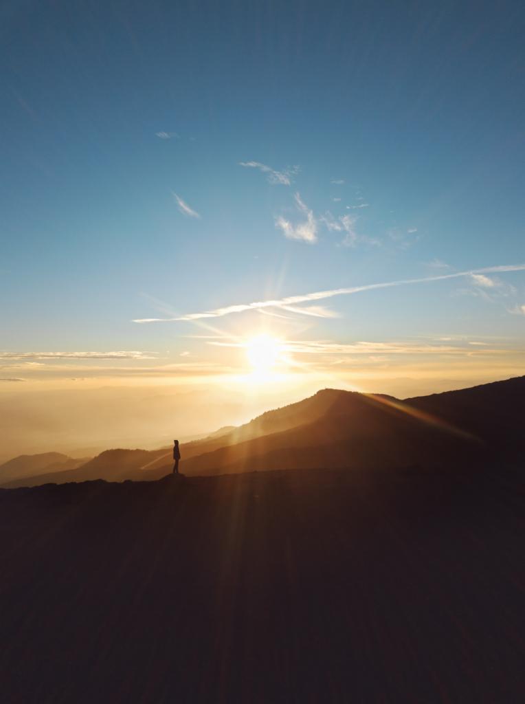 Sylwetka osoby stojącej na szczycie góry podczas wschodu słońca, z promieniami światła przebijającymi się przez mgłę i chmury.