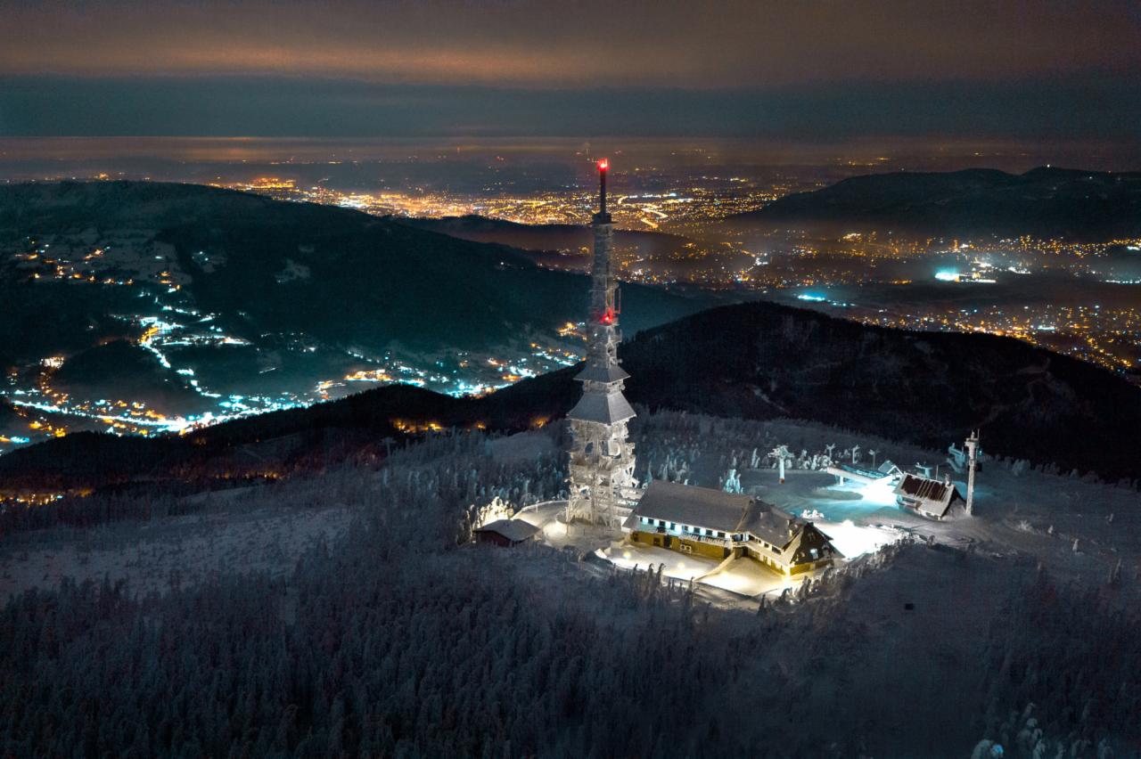 Nocny widok z lotu ptaka na ośnieżony szczyt góry ze stacją przekaźnikową i budynkami, w tle rozświetlone miasto.