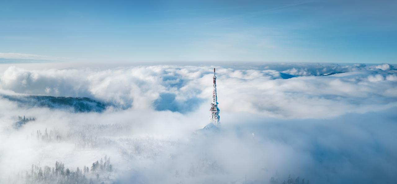 Zimowy krajobraz z wieżą przekaźnikową wystającą z morza chmur, pokrytą szronem, widok z lotu ptaka w słoneczny dzień.