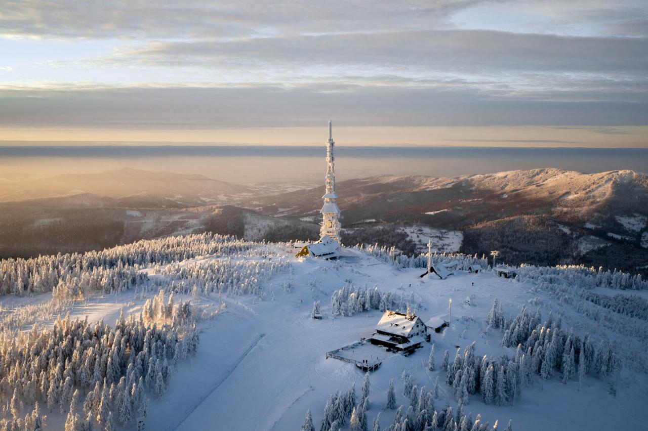 Zimowy krajobraz górski z wieżą przekaźnikową pokrytą szronem, widok z lotu ptaka na zaśnieżone drzewa i budynki, mglista dolina w tle.