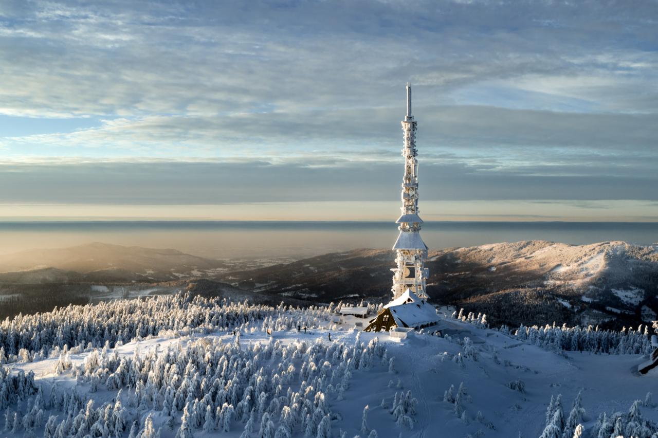 Zimowy krajobraz górski z wieżą telekomunikacyjną pokrytą śniegiem, widoczną na tle zachmurzonego nieba i zalesionych wzgórz.