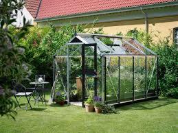 Szklarnia z czarną ramą na trawniku przed domem z czerwoną dachówką, obok stolik z laptopem i krzesłem.