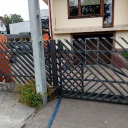 Nowoczesna, metalowa konstrukcja z ukośnymi prętami tworząca wzór rombów, z przesuwną bramą garażową w tle i fragmentem pomarańczowej dekoracji ogrodowej po lewej stronie.