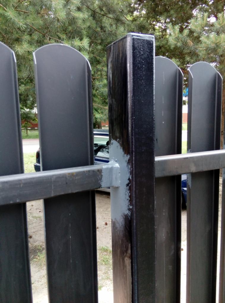 Szczegół metalowej, pomalowanej na czarno konstrukcji ogrodzenia panelowego z widocznymi śladami szarej farby na łączeniach, na tle zielonych drzew i fragmentu samochodu.