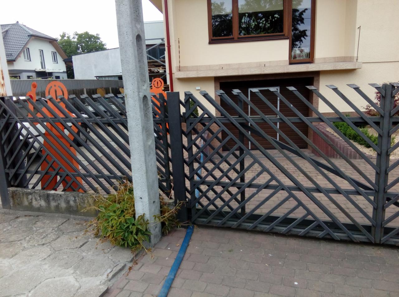 Nowoczesna, metalowa konstrukcja z ukośnymi prętami tworząca wzór rombów, z przesuwną bramą garażową w tle i fragmentem pomarańczowej dekoracji ogrodowej po lewej stronie.