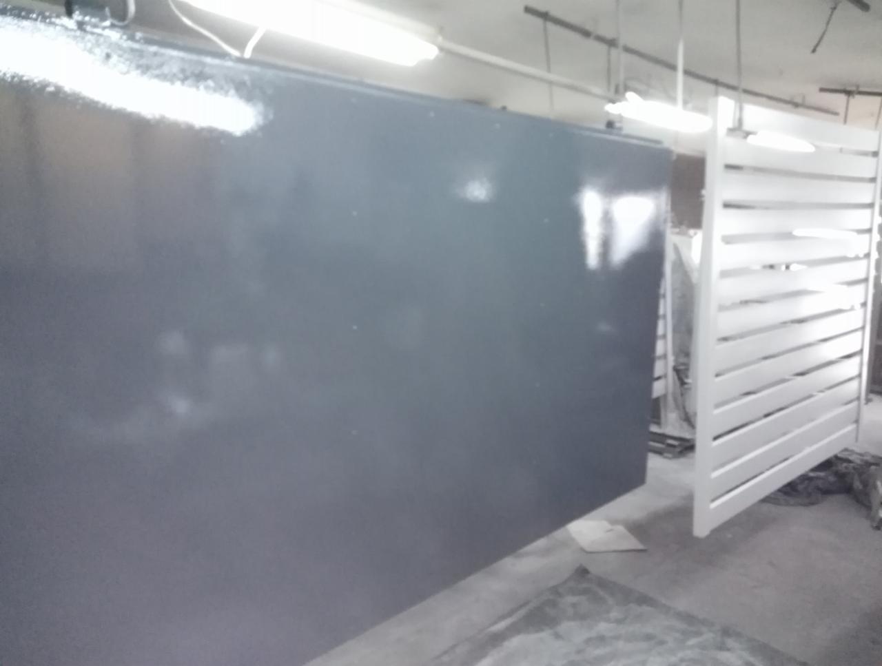 Elementy metalowe, prawdopodobnie fragmenty bramy, świeżo po malowaniu proszkowym w hali produkcyjnej, widoczne ślady farby w powietrzu.