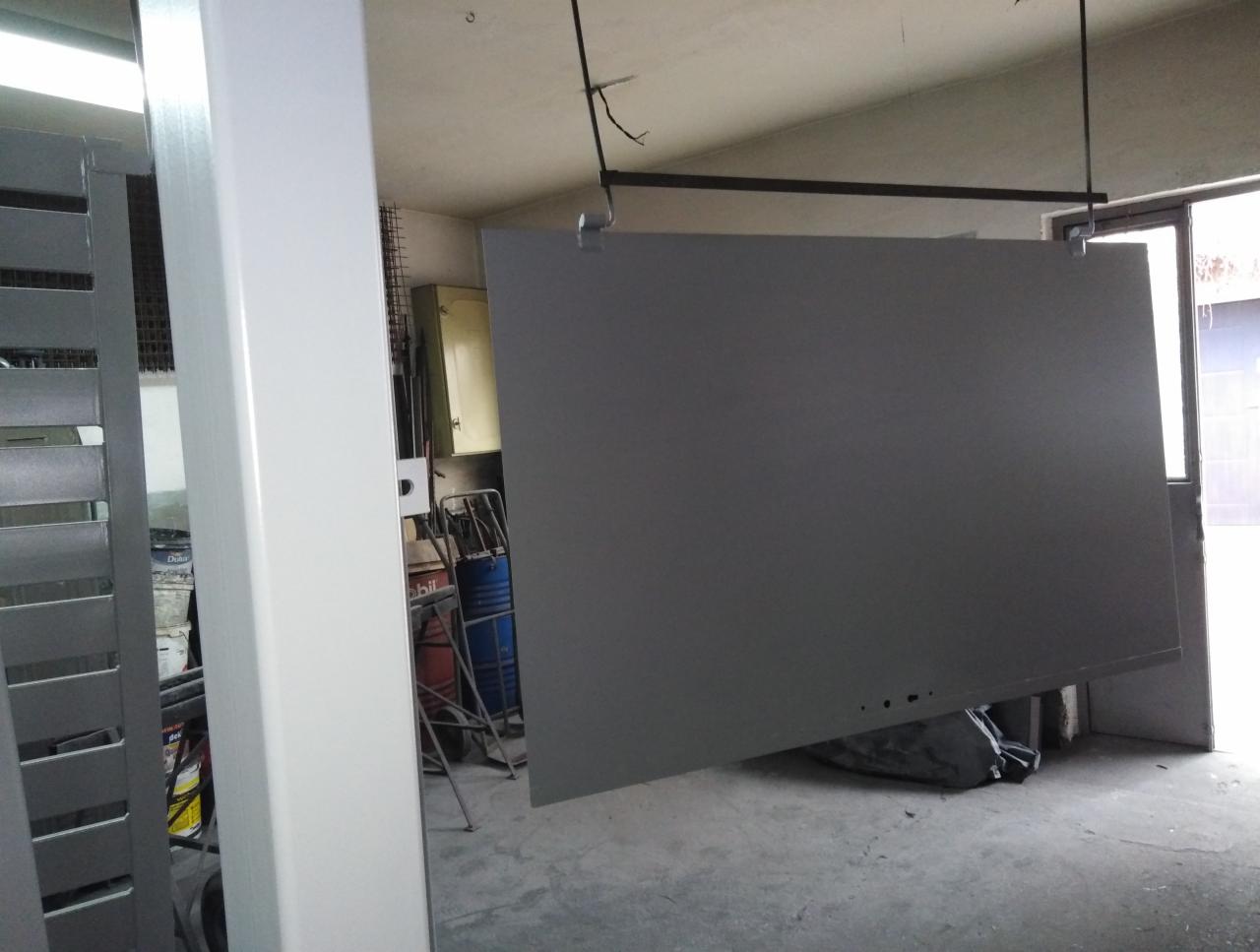 Szary panel bramy wjazdowej zawieszony na haku w warsztacie, widoczna część pomalowanej ramy bramy oraz wyposażenie warsztatu w tle.