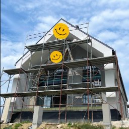 Firma Remontowo-budowlana B-Art - Dom w trakcie ocieplania elewacji, widoczne rusztowania, częściowo otynkowana ściana i fragment fundamentu z izolacją przeciwwilgociową, na rusztowaniu stoi pracownik.