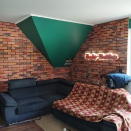 Firma Remontowo-budowlana B-Art - Pokój po remoncie: ceglana ściana z podświetlanym napisem Harley-Davidson, ciemna sofa z kocem w kratę, pomalowany na zielono fragment ściany pod sufitem, widoczny mężczyzna pochylony nad czymś...