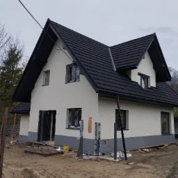 Niedawno otynkowany dom jednorodzinny z ciemną dachówką i szarym cokołem, widoczne elementy rusztowania, teren wokół domu w trakcie prac wykończeniowych, góry w tle.