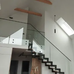 Nowoczesne schody ażurowe z drewnianymi stopniami i szklaną balustradą, widoczne drewniane belki pod sufitem, w tle drabina malarska i akcesoria remontowe.