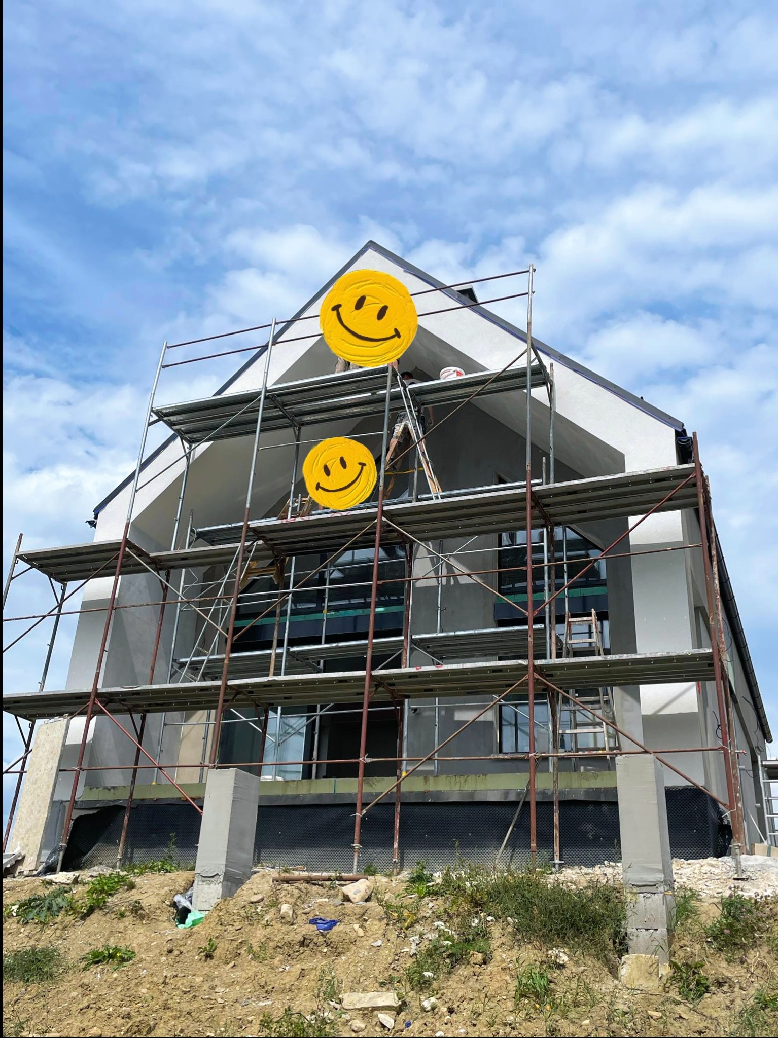 Dom w trakcie ocieplania elewacji, widoczne rusztowania, częściowo otynkowana ściana i fragment fundamentu z izolacją przeciwwilgociową, na rusztowaniu stoi pracownik.