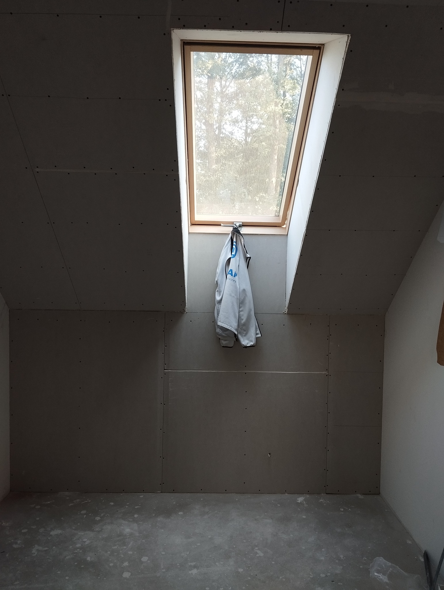 Poddasze w trakcie wykańczania płytami karton gips, widoczne łączenia płyt i wkręty, okno dachowe z powieszonym ubraniem roboczym.