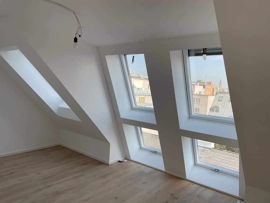 Poddasze z trzema oknami dachowymi, świeżo pomalowane na biało, z drewnianą podłogą i wiszącą żarówką na kablu.