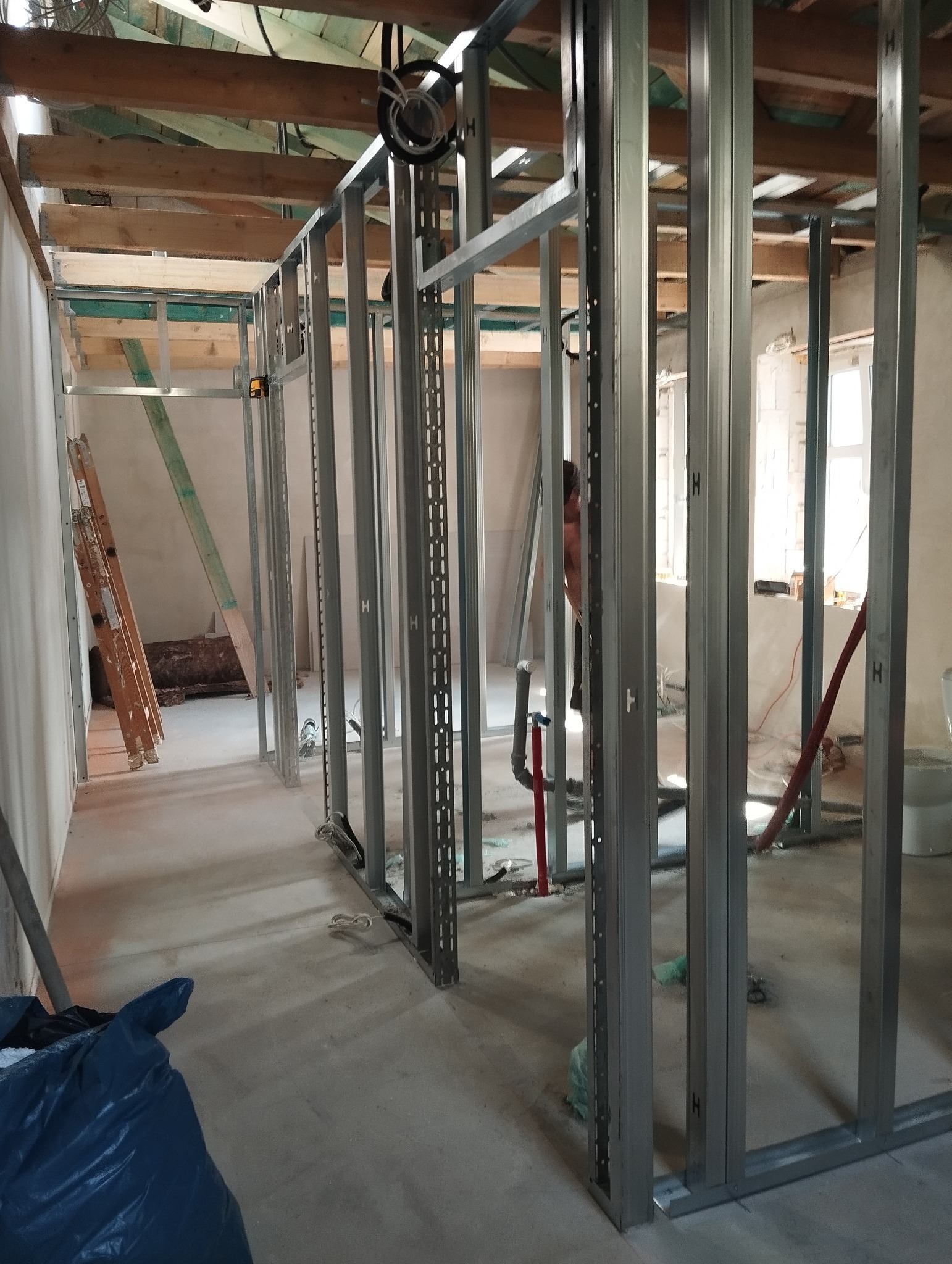 Metalowa konstrukcja szkieletowa ścian działowych w surowym stanie, widoczne profile z otworami, drewniane belki stropowe, instalacje elektryczne i sanitarne w tle, przygotowanie do montażu okładzin.