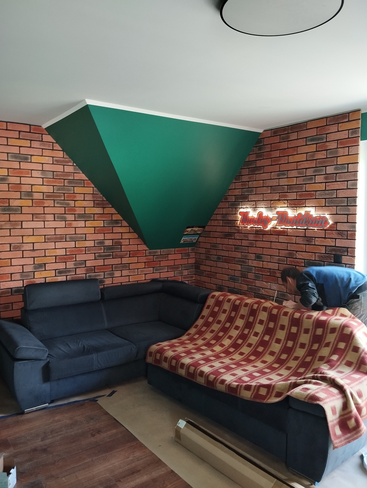 Pokój po remoncie: ceglana ściana z podświetlanym napisem Harley-Davidson, ciemna sofa z kocem w kratę, pomalowany na zielono fragment ściany pod sufitem, widoczny mężczyzna pochylony nad czymś...