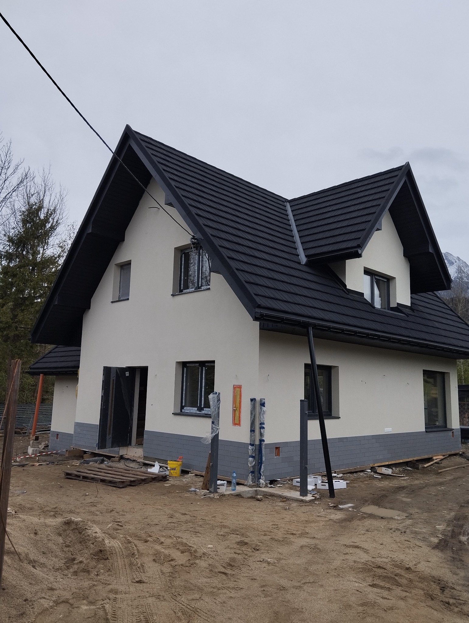 Niedawno otynkowany dom jednorodzinny z ciemną dachówką i szarym cokołem, widoczne elementy rusztowania, teren wokół domu w trakcie prac wykończeniowych, góry w tle.