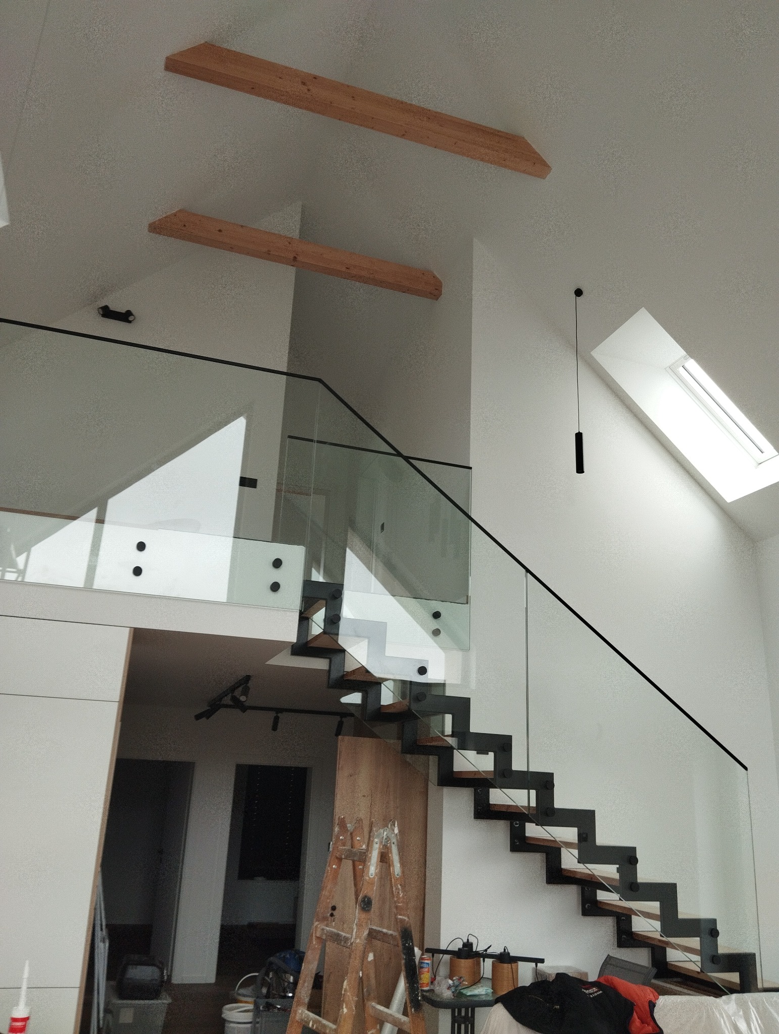 Nowoczesne schody ażurowe z drewnianymi stopniami i szklaną balustradą, widoczne drewniane belki pod sufitem, w tle drabina malarska i akcesoria remontowe.
