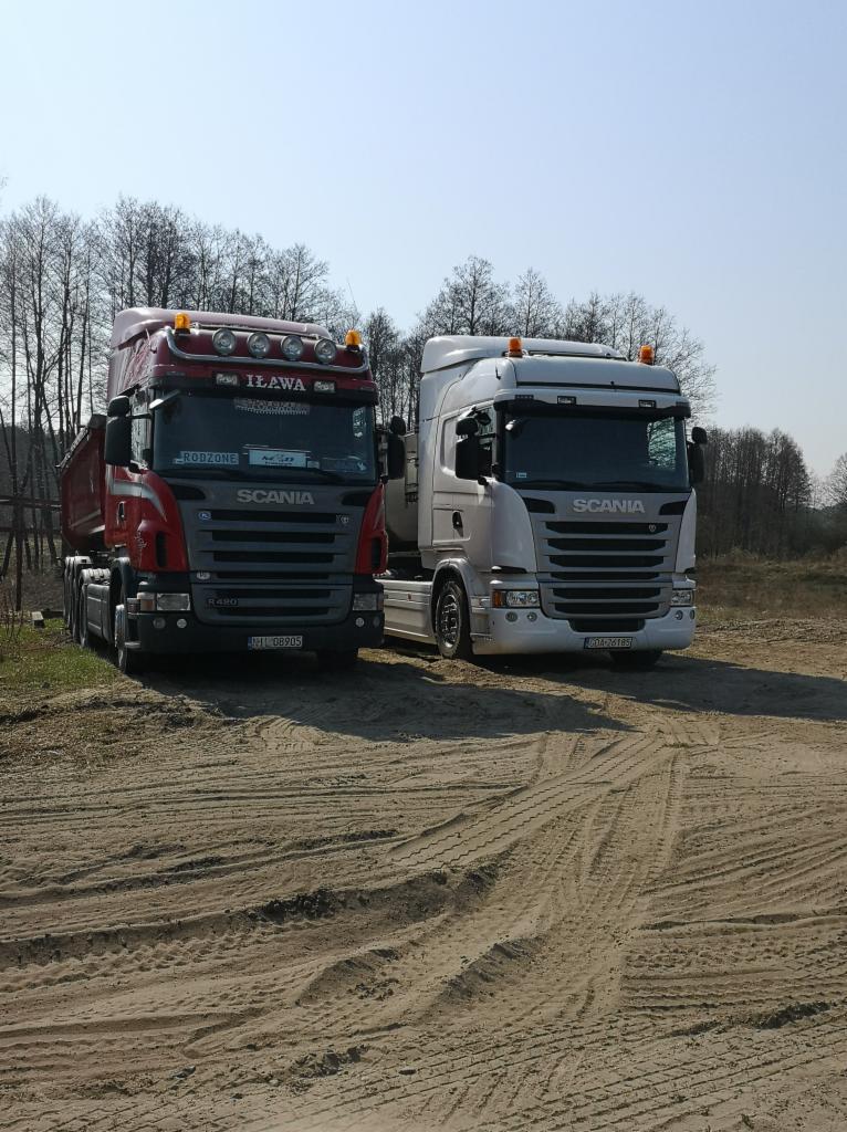Dwa samochody ciężarowe marki Scania, czerwony i biały, zaparkowane na piaszczystym placu z widocznymi śladami opon, w tle drzewa.