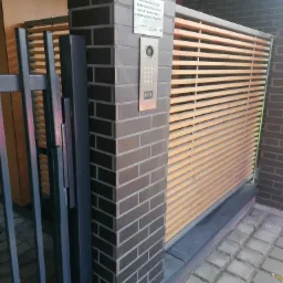 Nowoczesna brama wjazdowa z drewnianymi lamelami, domofon przy słupku z ciemnej cegły, kostka brukowa na podjeździe.