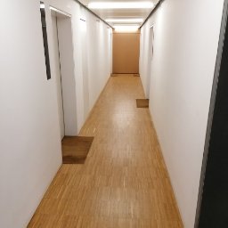 AGRA - Długi, pusty korytarz biurowy z drewnianą podłogą i białymi ścianami, oświetlony przez lampy sufitowe, z widocznymi drzwiami i wycieraczkami.