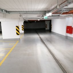 AGRA - Wnętrze czystego, jasnego parkingu podziemnego z żółtymi liniami na szarej posadzce i oznaczeniami ostrzegawczymi na filarach.