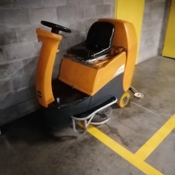 AGRA - Pomarańczowo-szara maszyna do czyszczenia podłóg z siedzeniem, stojąca na betonowej posadzce obok żółtej linii w hali.