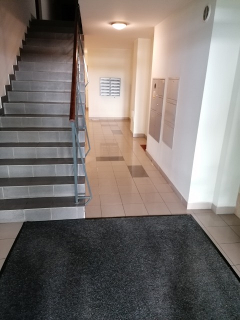 Wnętrze klatki schodowej w budynku biurowym z szarymi schodami, beżową podłogą z płytek i ciemnoszarą wycieraczką przy wejściu.