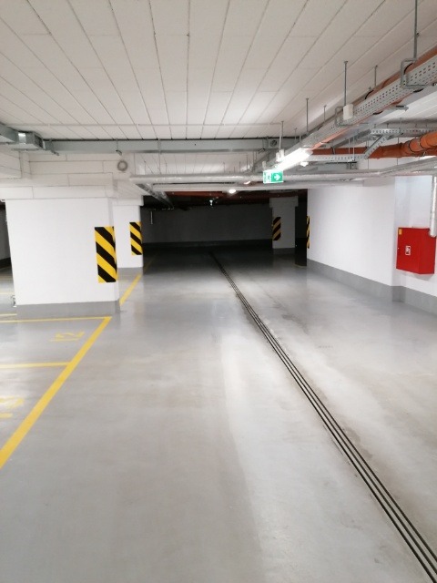Wnętrze czystego, jasnego parkingu podziemnego z żółtymi liniami na szarej posadzce i oznaczeniami ostrzegawczymi na filarach.