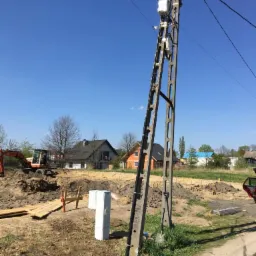 Wykonanie wykopu pod fundamenty domu jednorodzinnego z widoczną skrzynką przyłączeniową i koparką w tle, słup energetyczny na pierwszym planie.