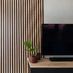 Panel ścienny z drewnianych listew w salonie, obok szafki RTV z czarnymi frontami i blatem imitującym drewno, na której stoi roślina doniczkowa i tablet.