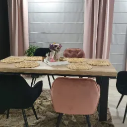 Jadalnia z drewnianym stołem, nakrytym sześcioma okrągłymi podkładkami, otoczona tapicerowanymi krzesłami w kolorze pudrowego różu i czerni na tle zasłon w odcieniach różu i szarości.