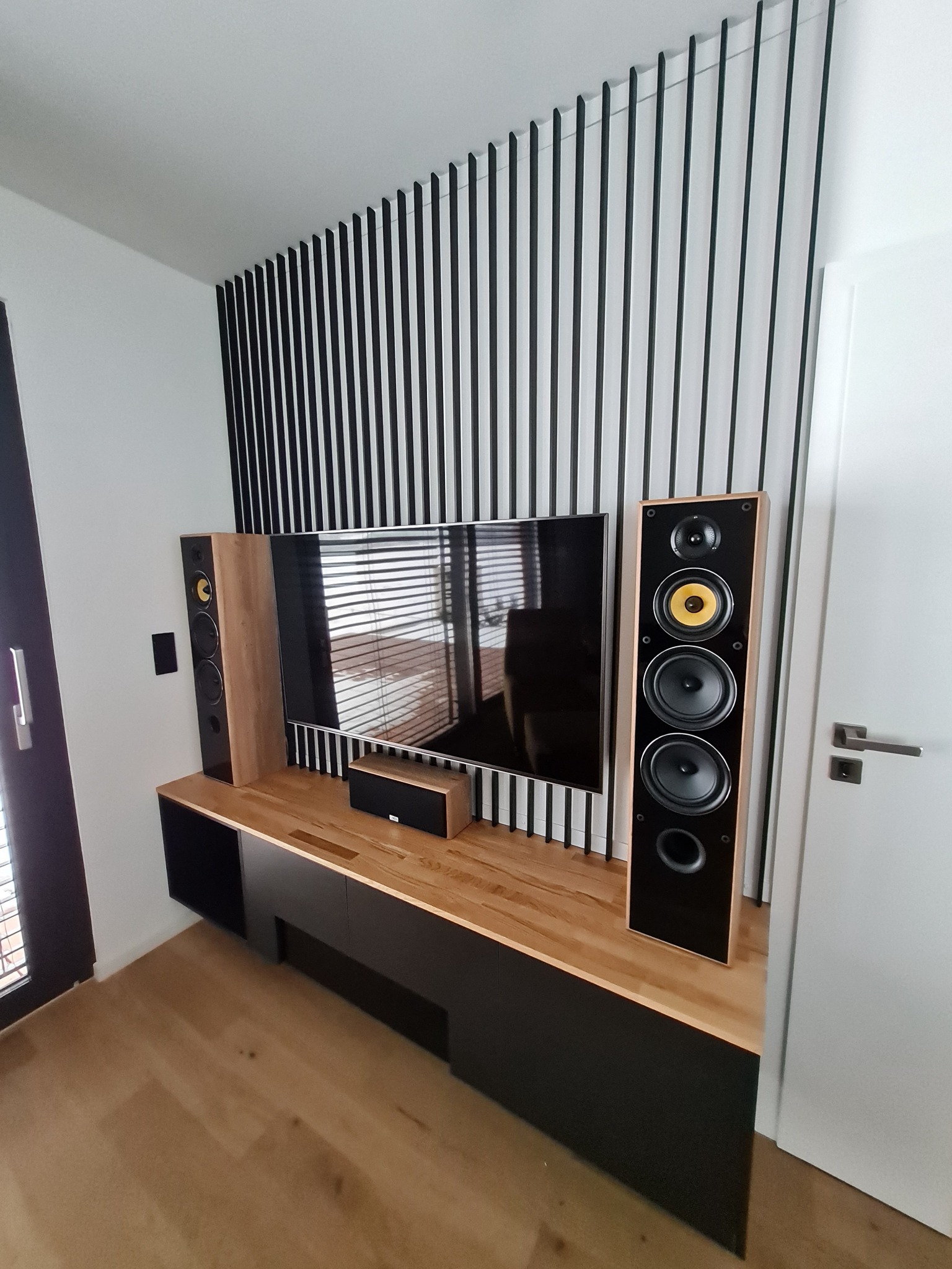 Nowoczesna aranżacja salonu z drewnianym blatem pod telewizor, czarnymi szafkami, głośnikami i dekoracyjną ścianką z pionowych listewek.