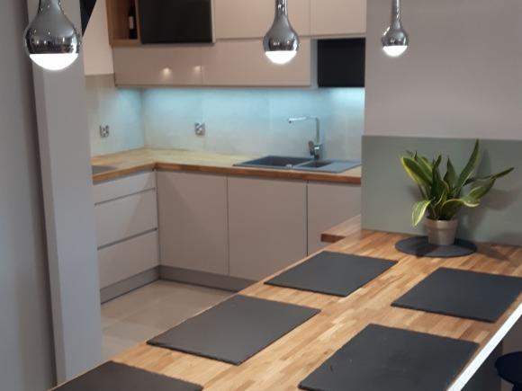 Nowoczesna kuchnia z drewnianym blatem i minimalistycznymi lampami wiszącymi, zlew wpuszczony w blat, roślina doniczkowa na blacie, szare podkładki pod talerze.