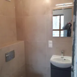 Wykończona łazienka z beżowymi płytkami imitującymi marmur, podwieszaną toaletą, umywalką z szafką w kolorze grafitowym i lustrem z oświetleniem LED. W lustrze odbija się osoba robiąca zdjęcie.