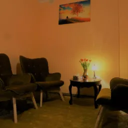 Pokój konsultacyjny z dwoma zielonymi fotelami, małym stolikiem z tulipanami, lampką i pudełkiem chusteczek, na ścianie obraz z drzewem.