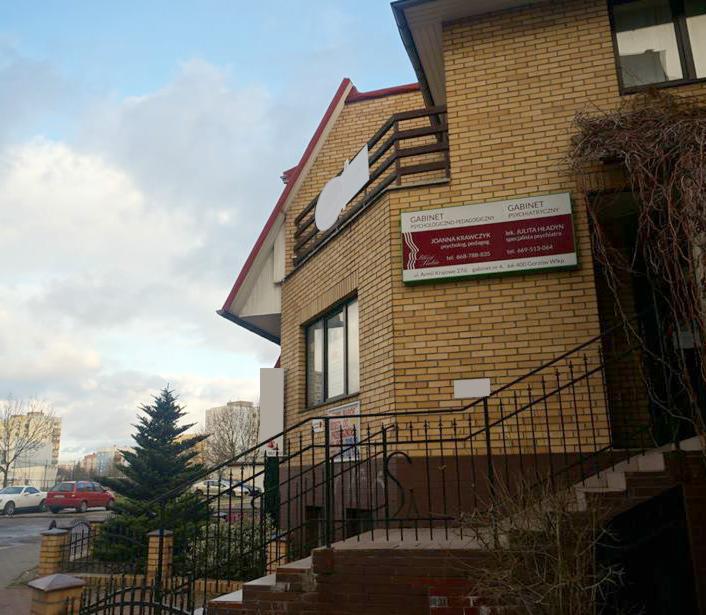 Budynek z cegły z szyldami gabinetów: psychologiczno-pedagogicznego Joanna Krawczyk i psychiatrycznego L. Julita Hładyn, widok z zewnątrz, schody wejściowe z metalową barierką.