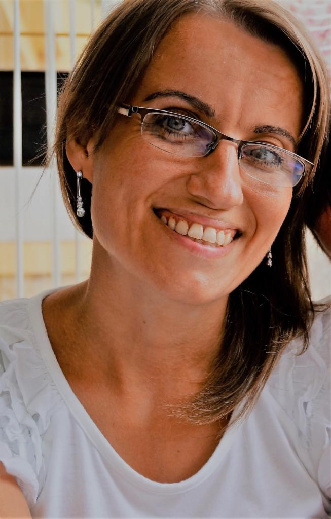 Portret uśmiechniętej kobiety w okularach i białej bluzce, z kolczykami, w jasnym wnętrzu.