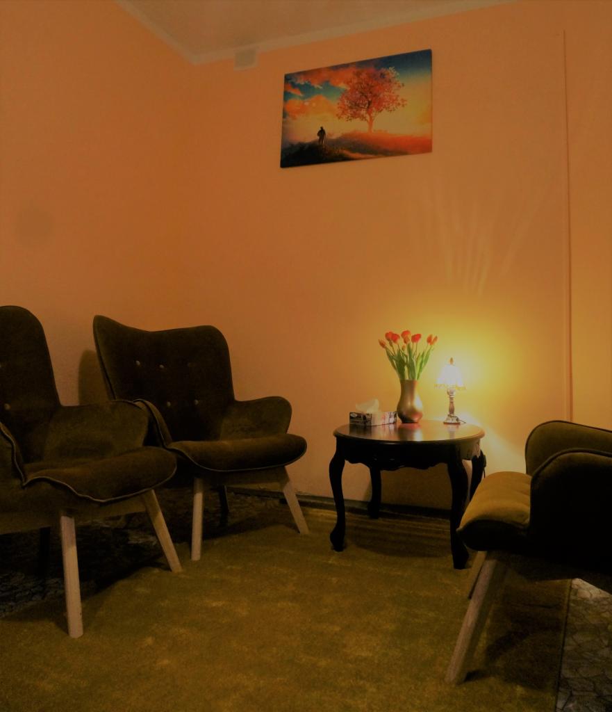 Pokój konsultacyjny z dwoma zielonymi fotelami, małym stolikiem z tulipanami, lampką i pudełkiem chusteczek, na ścianie obraz z drzewem.